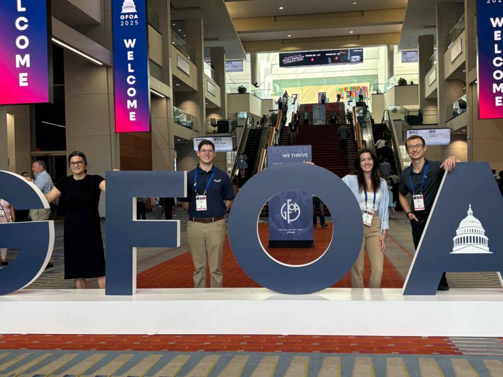 Four PayIt employees standing and smiling behind large 'GFOA' letters at the GFOA 2025 conference, with welcome banners and an escalator in the background.
